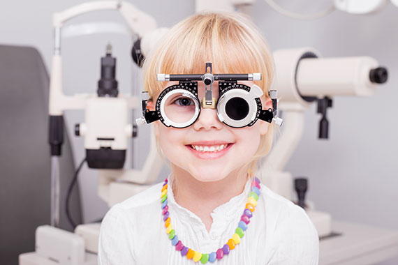 Ein junges Mädchen sitzt in einem Augenuntersuchungsraum und schaut neugierig und erwartungsvoll auf eine Augentabelle.